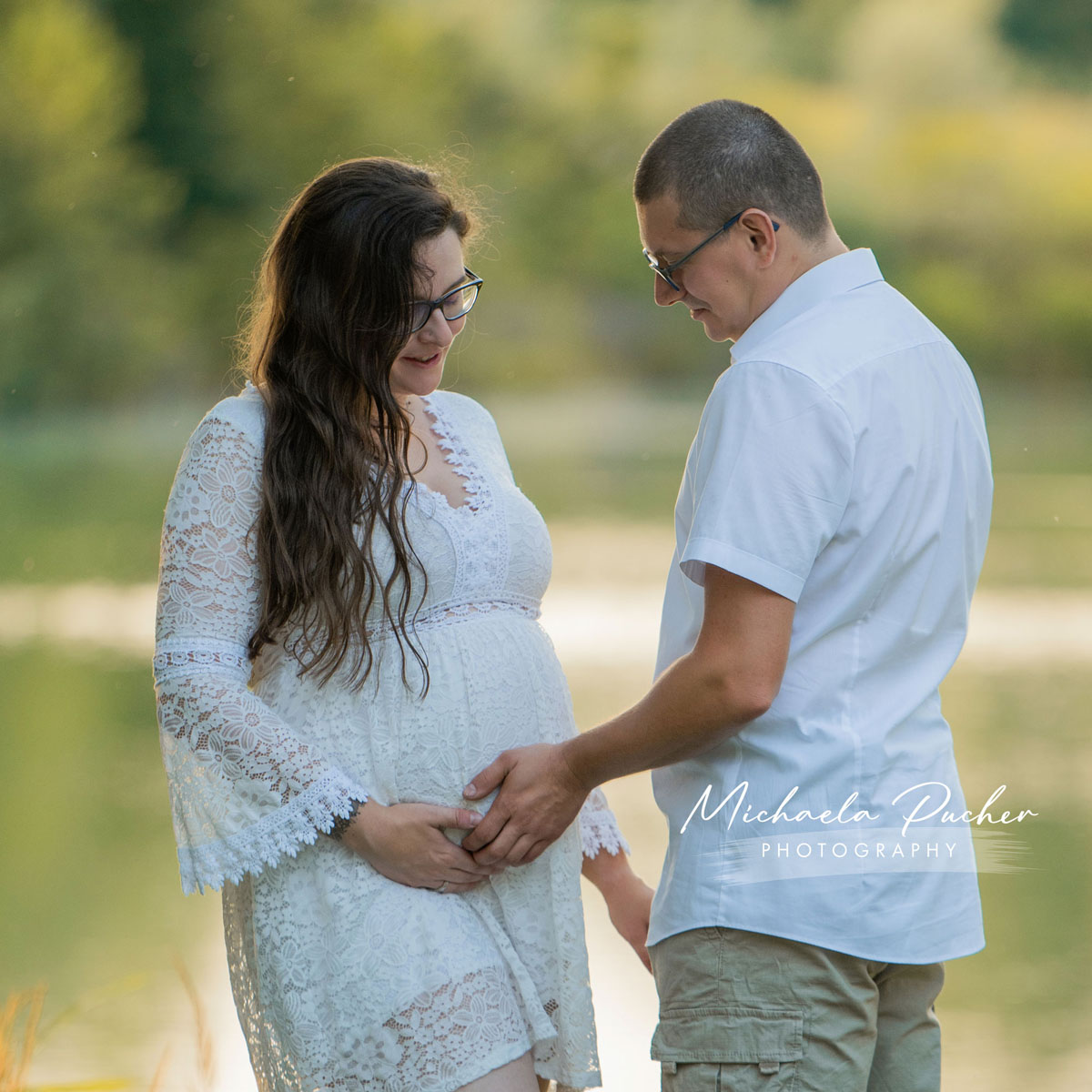 Schwangere Frau in weißem Spitzenkleid steht am Ufer mit ihrem Partner, beide halten liebevoll den Babybauch und schauen lächelnd darauf.